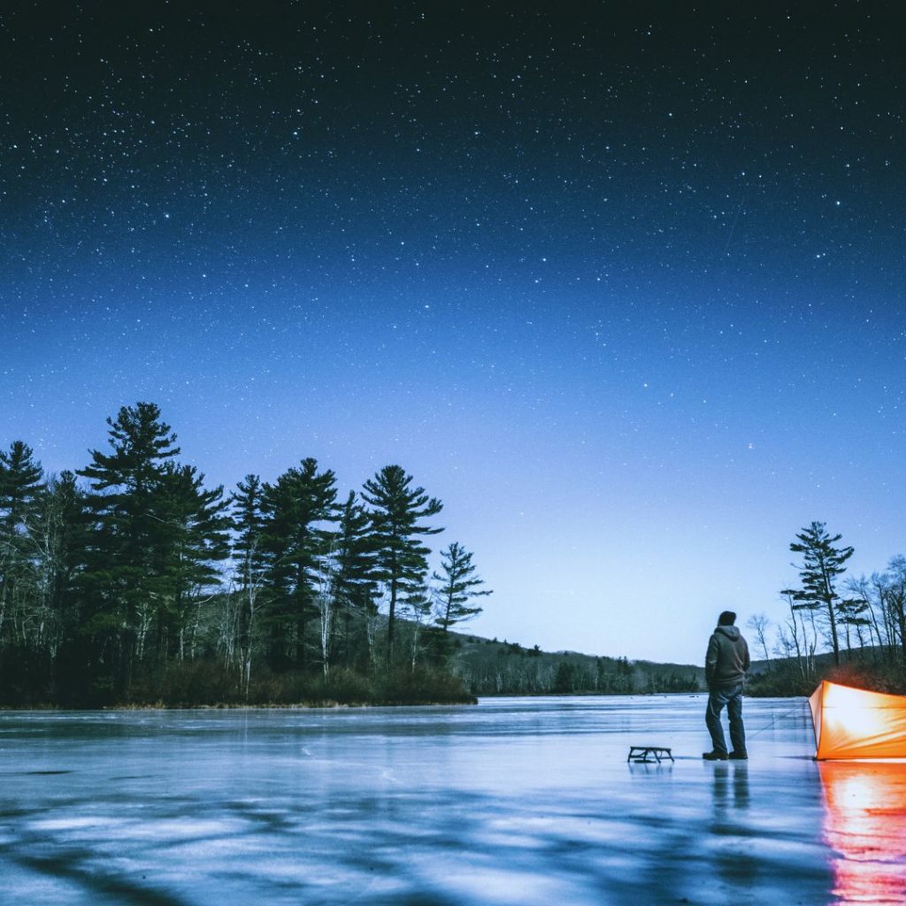 Stargazing man on a frozen lake with pine trees and stars overhead